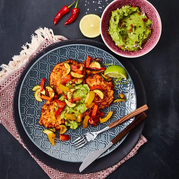 Süßkartoffel-Rösti mit würzigem Fajita-Gemüse aus Paprika, Zucchini und Tomaten, serviert auf einem orientalischen Teller. Beilage: cremige Guacamole in einer Schale, garniert mit frischer Limette und roten Chilischoten. Dekoriert mit einem gemusterten Serviertuch und stilvollem Holz-Besteck.