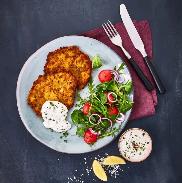 Zwei goldbraun gebratene Gemüsetaler, serviert auf einem Teller mit Kresse-Kräuterquark und einem frischen Salat aus Feldsalat, Radieschen, roten Zwiebeln und einer leichten Vinaigrette. Daneben liegt ein kleines Schälchen mit Quarkdip sowie Zitronenspalten, angerichtet auf einem dunklen Hintergrund mit Besteck auf einer weinroten Stoffserviette.