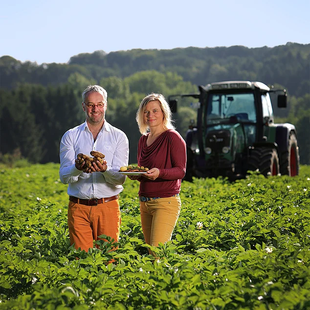Uwe und Marion Pahmeyer stehen inmitten eines grünen Kartoffelfeldes und lächeln in die Kamera. Uwe hält frische, erdige Kartoffeln in den Händen, während Marion einen Teller mit geschälten Kartoffeln präsentiert. Im Hintergrund ist ein Traktor leicht unscharf erkennbar, und die Umgebung zeigt eine grüne, ländliche Landschaft.