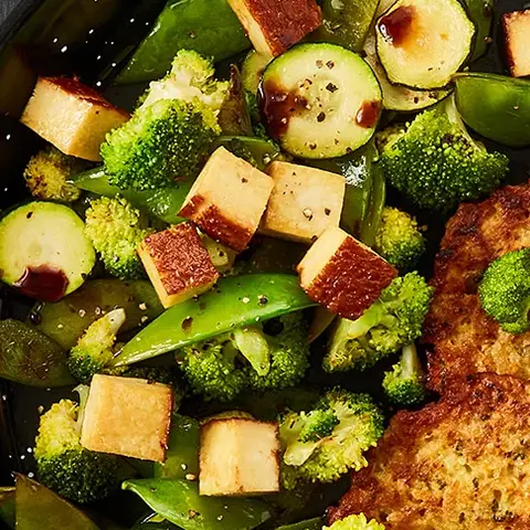 Zwei goldbraune Gemüsetaler, serviert in einer schwarzen Pfanne mit einer Gemüse-Tofu-Mischung aus Zucchini, Brokkoli, Zuckerschoten und knusprig angebratenen Tofuwürfeln, garniert mit dunkler Soße. Neben der Pfanne liegen eine Metall-Bratwender, grobes Meersalz und Pfefferkörner in kleinen Schalen auf einem dunklen Hintergrund.