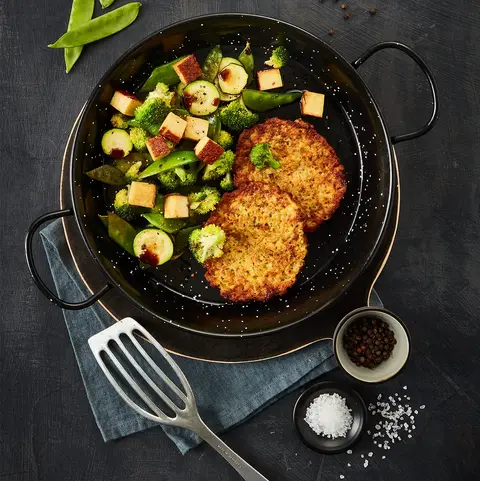 Zwei goldbraune Gemüsetaler, serviert in einer schwarzen Pfanne mit einer Gemüse-Tofu-Mischung aus Zucchini, Brokkoli, Zuckerschoten und knusprig angebratenen Tofuwürfeln, garniert mit dunkler Soße. Neben der Pfanne liegen eine Metall-Bratwender, grobes Meersalz und Pfefferkörner in kleinen Schalen auf einem dunklen Hintergrund.