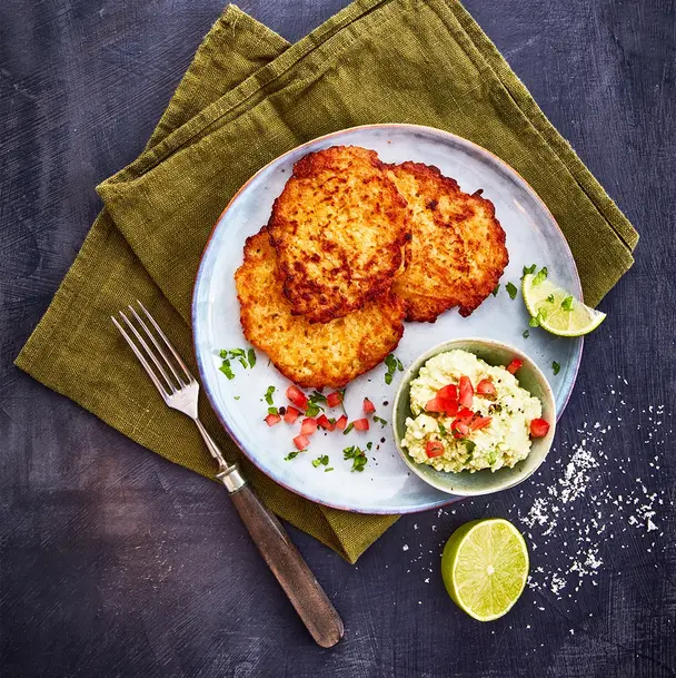 Ein Teller mit drei knusprigen Reibekuchen, serviert mit einer kleinen Schale Avocadocreme, garniert mit gehackten Tomaten und frischen Kräutern. Eine halbe Limette und grobes Meersalz liegen dekorativ daneben. Das Arrangement ist auf einer olivgrünen Stoffserviette präsentiert, begleitet von einer Gabel mit Holzgriff, alles auf einem dunklen Hintergrund.