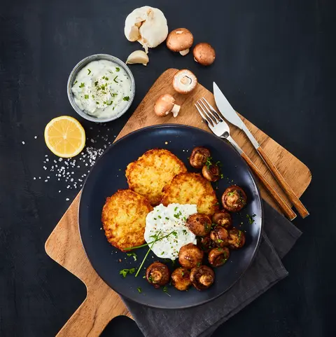 Ein Teller mit drei knusprigen Reibekuchen, serviert mit gebratenen Champignons und einem Kräuterdip, garniert mit frischen Kräutern. Das Arrangement steht auf einem Holzbrett, begleitet von einer halben Zitrone, einer Schale mit Kräuterdip, Knoblauchknollen und frischen Champignons. Besteck mit Holzgriffen liegt dekorativ daneben, alles auf einem dunklen Untergrund.