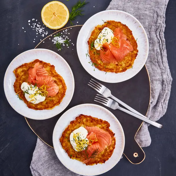 Drei Teller mit knusprigen Reibekuchen, belegt mit Räucherlachs, einem Klecks Sauerrahm und garniert mit Dill und Zitronenzesten. Die Teller sind auf einem dunklen Untergrund angerichtet, begleitet von einer halben Zitrone, frischen Dillzweigen und grobem Meersalz. Besteck liegt dekorativ auf einer grauen Stoffserviette.