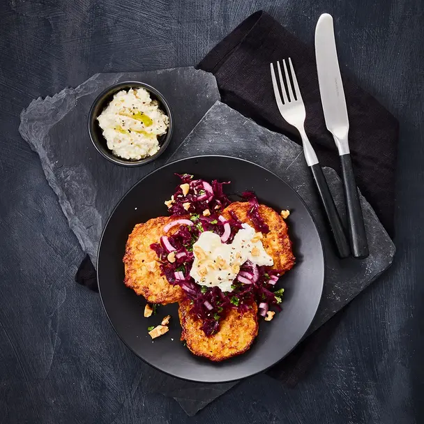Drei knusprige Süßkartoffel-Rösti auf einem schwarzen Teller, garniert mit würzigem Rotkrautsalat, zerbröckeltem Fetakäse und gehackten Nüssen. Die dunkle Schieferplatte und das stilvolle Besteck runden die edle Präsentation ab. Eine kleine Schale mit cremigem Feta-Dip mit Olivenöl ergänzt das Gericht. 