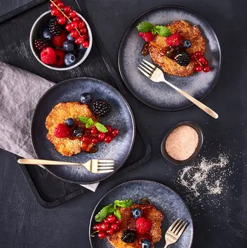 Drei Teller mit Mini-Reibekuchen, bestreut mit Zimtzucker, belegt mit einer Auswahl frischer Beeren wie Himbeeren, Brombeeren, Johannisbeeren und Heidelbeeren, garniert mit Minzblättern. Darüber wurde Sirup gegossen. Daneben steht eine Schale mit Beeren als Dekoration sowie eine kleine Schale mit Zimtzucker, alles auf einem dunklen Hintergrund mit goldfarbenen Dessertgabeln arrangiert.