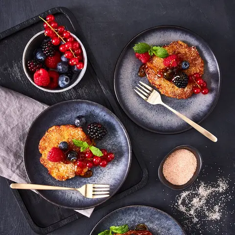 Drei Teller mit Mini-Reibekuchen, bestreut mit Zimtzucker, belegt mit einer Auswahl frischer Beeren wie Himbeeren, Brombeeren, Johannisbeeren und Heidelbeeren, garniert mit Minzblättern. Darüber wurde Sirup gegossen. Daneben steht eine Schale mit Beeren als Dekoration sowie eine kleine Schale mit Zimtzucker, alles auf einem dunklen Hintergrund mit goldfarbenen Dessertgabeln arrangiert.