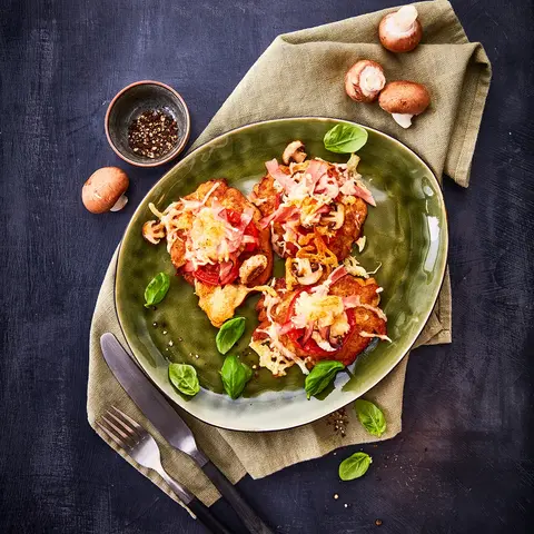 Krosse Rösti-Pizza mit Tomatenscheiben, frischen Champignons, Schinken und geschmolzenem Käse, serviert auf einem grünen Teller, dekoriert mit Basilikumblättern und begleitet von frischen Champignons sowie einer Pfeffer-Mühle im Hintergrund.