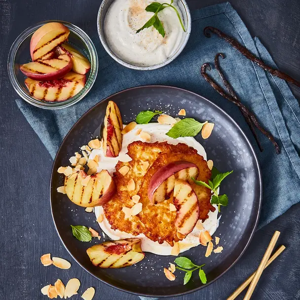 Knusprige Rösti serviert mit cremigem Vanillequark, gegrillten Nektarinenspalten, gerösteten Mandeln und frischen Minzblättern auf einem dunklen Teller. Daneben Schalen mit weiteren Zutaten wie Nektarinen und Vanillequark, dekoriert mit Vanilleschoten und einem blauen Stoffservietten.