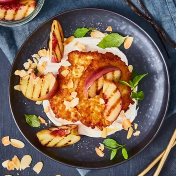 Knusprige Rösti serviert mit cremigem Vanillequark, gegrillten Nektarinenspalten, gerösteten Mandeln und frischen Minzblättern auf einem dunklen Teller. Daneben Schalen mit weiteren Zutaten wie Nektarinen und Vanillequark, dekoriert mit Vanilleschoten und einem blauen Stoffservietten.