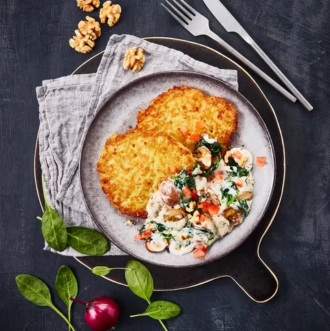 Reibekuchen serviert mit einer cremigen Gorgonzola-Sauce, Spinat und Champignons, dekoriert mit Walnüssen auf dunklem Untergrund.