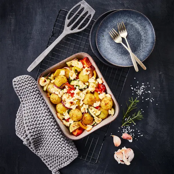 Rosmarinkartoffeln mit Ofengemüse wie Zucchini, Paprika und roten Zwiebeln, überbacken mit zerbröseltem Feta-Käse, serviert in einer Backform auf einem schwarzen Gitter. Das Gericht wird von einer grauen Topflappen-Dekoration, Rosmarinzweig, Knoblauchzehen und grobem Meersalz ergänzt. Zwei moderne Teller mit goldenen Gabeln stehen bereit.