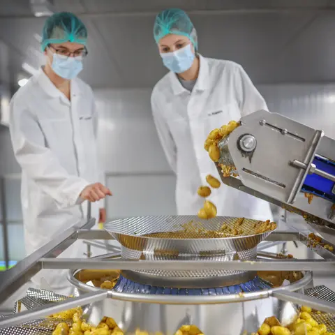 Mitarbeiter in Schutzkleidung bei der Arbeit an einer modernen Mehrkopfwaage, die marinierte Kartoffeln portioniert und verteilt.