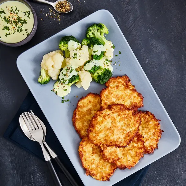 Krosse Rösti, serviert mit gedünstetem Blumenkohl und Brokkoli, garniert mit cremiger Sauce und Kräutern auf einem blauen Teller. Im Hintergrund frische Gewürze und eine passende Sauce im Schälchen.