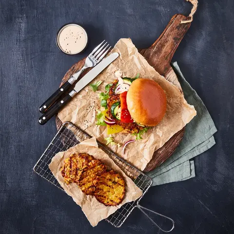 Ein vegetarischer Burger mit Gemüsegrillis als Patty, frischem Salat, Gurken, Tomaten und Zwiebeln, serviert auf Backpapier und einem rustikalen Holzbrett. Daneben liegt ein Gitter mit weiteren gegrillten Gemüsegrillis sowie ein kleines Schälchen mit Dip. Besteck befindet sich ebenfalls auf dem Holzbrett, alles auf einem dunklen Hintergrund.